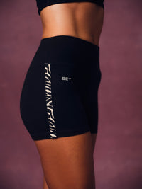 Black athletic shorts with a white vertical side stripe and 'SET' printed in white on the hip, shown on a model against a dusty mauve background. 