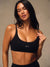A woman modeling a black athletic sports bra with a scoop neckline and the brand name 'SET' printed in white on the front, set against a neutral textured beige background. <model-info>Jelene wears a size XS and is 5’7”, 23” waist, 32AB bust.</model-info>