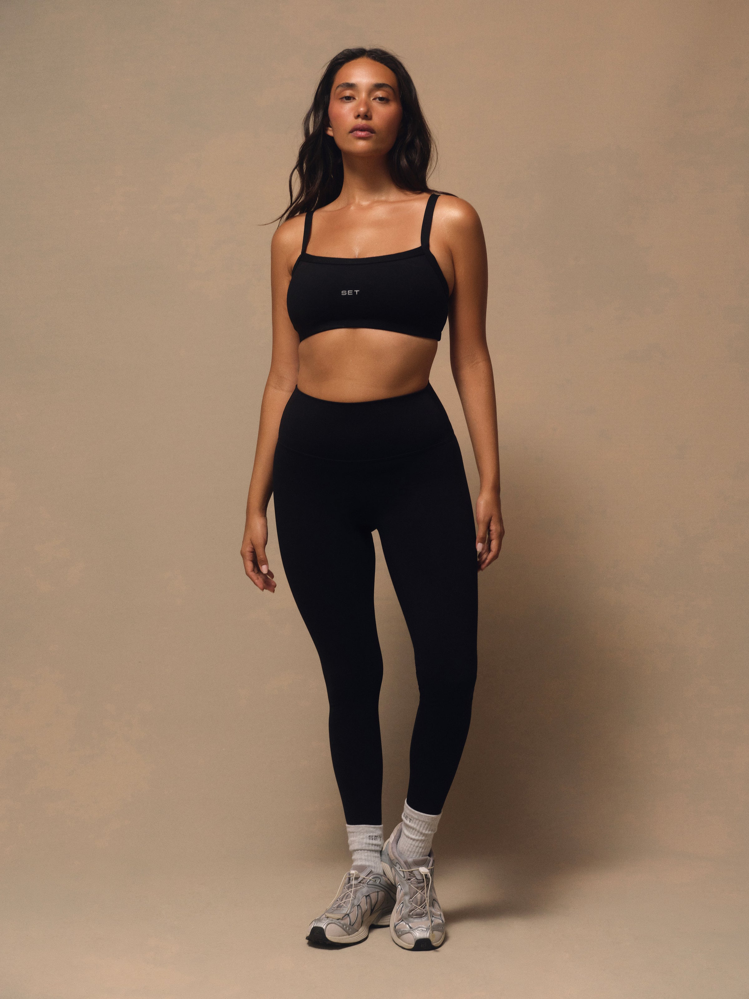 A woman modeling a black activewear set consisting of a square-neck sports bra and high-waisted leggings, paired with gray and white sneakers, against a textured beige studio backdrop. 