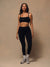 A woman modeling a black activewear set consisting of a square-neck sports bra and high-waisted leggings, paired with gray and white sneakers, against a textured beige studio backdrop. <model-info>Irene wears a size M and is 5’8”, 30” waist, 36" bust.</model-info>