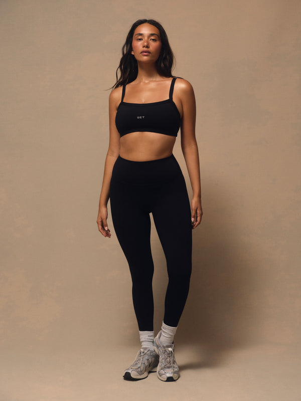A woman modeling a black activewear set consisting of a square-neck sports bra and high-waisted leggings, paired with gray and white sneakers, against a textured beige studio backdrop. 