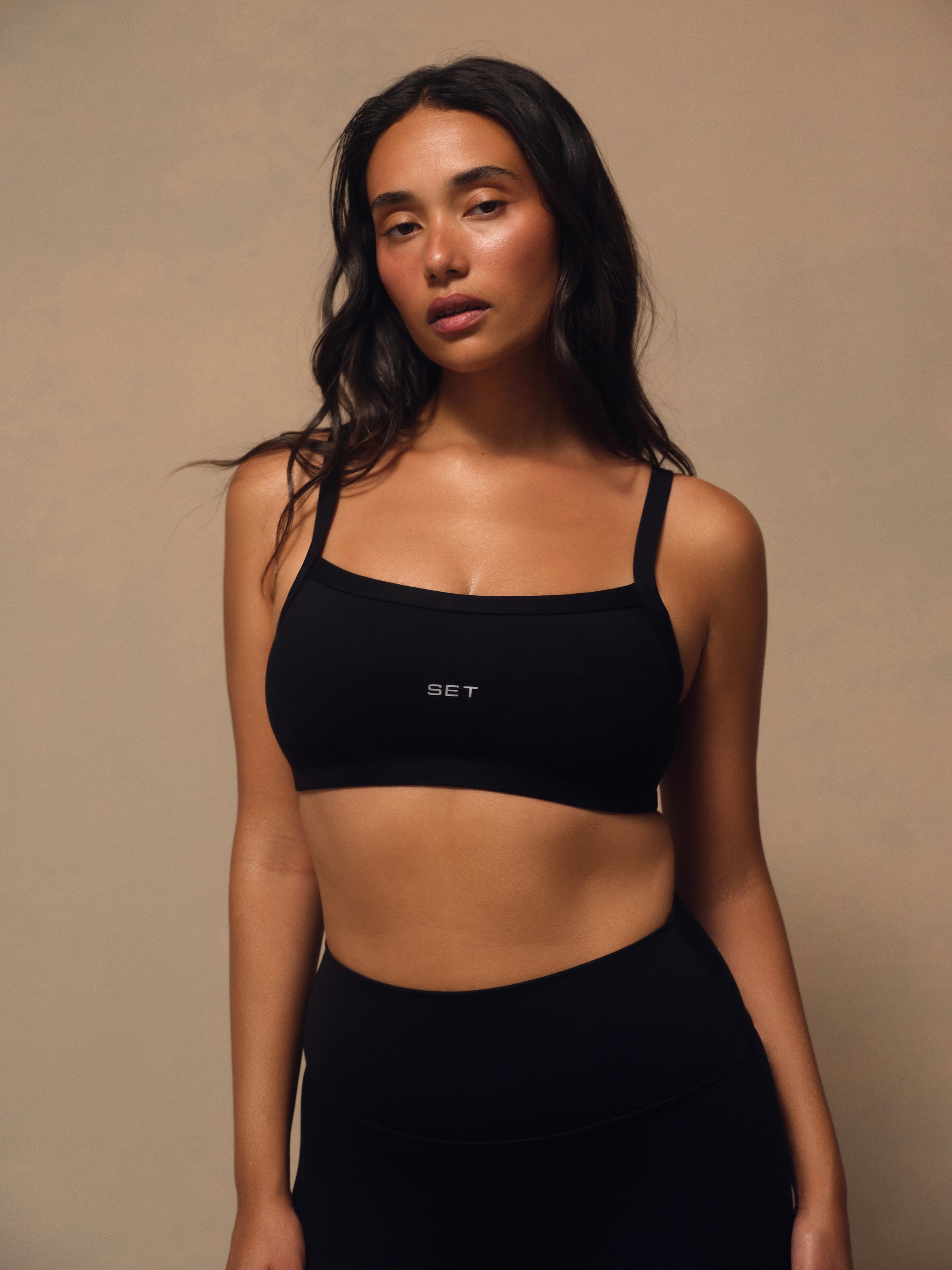 A woman modeling a black athletic sports bra with a square neckline and thin straps, featuring a small white 'SET' logo on the center chest, paired with black athletic bottoms against a warm beige studio background. 