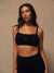A woman modeling a black athletic sports bra with a square neckline and thin straps, featuring a small white 'SET' logo on the center chest, paired with black athletic bottoms against a warm beige studio background. <model-info>Irene wears a size M and is 5’8”, 30” waist, 36" bust.</model-info>