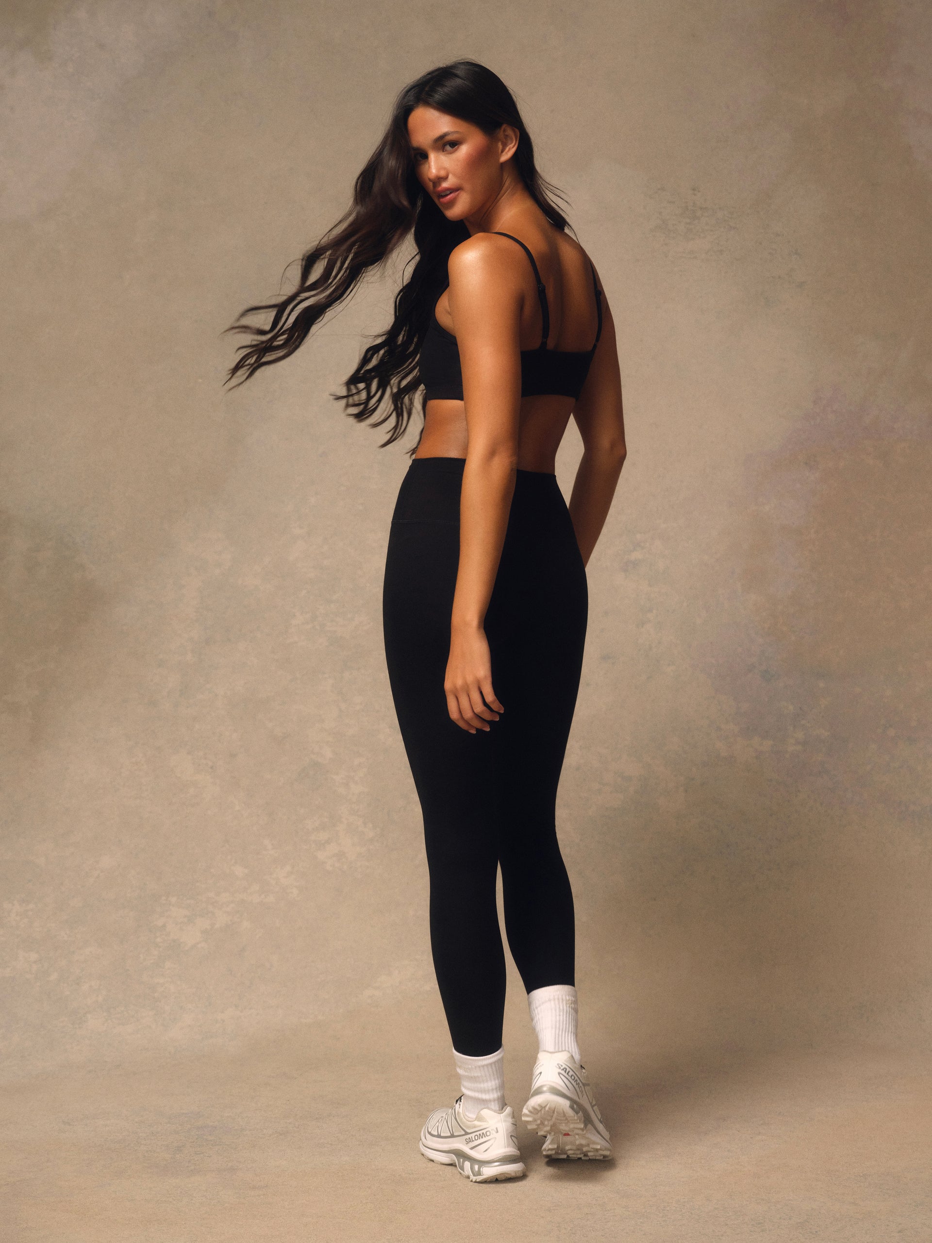 A woman posing in a black sports bra and black leggings with white socks and white sneakers, set against a textured beige studio backdrop.