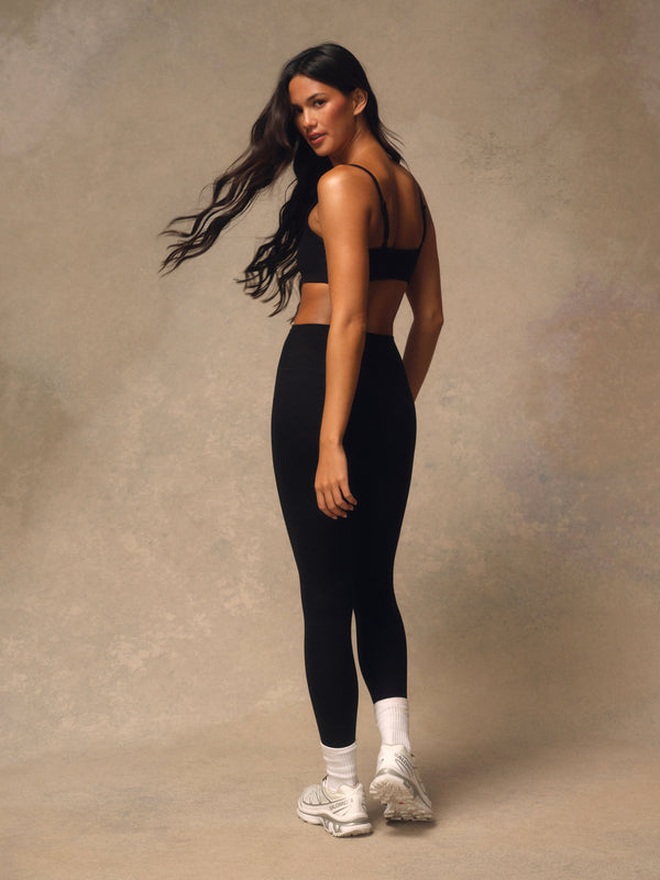A woman posing in a black sports bra and black leggings with white socks and white sneakers, set against a textured beige studio backdrop.