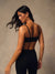 A woman with long dark hair is shown from the back wearing a black sports bra and black high-waisted leggings against a textured beige studio backdrop.