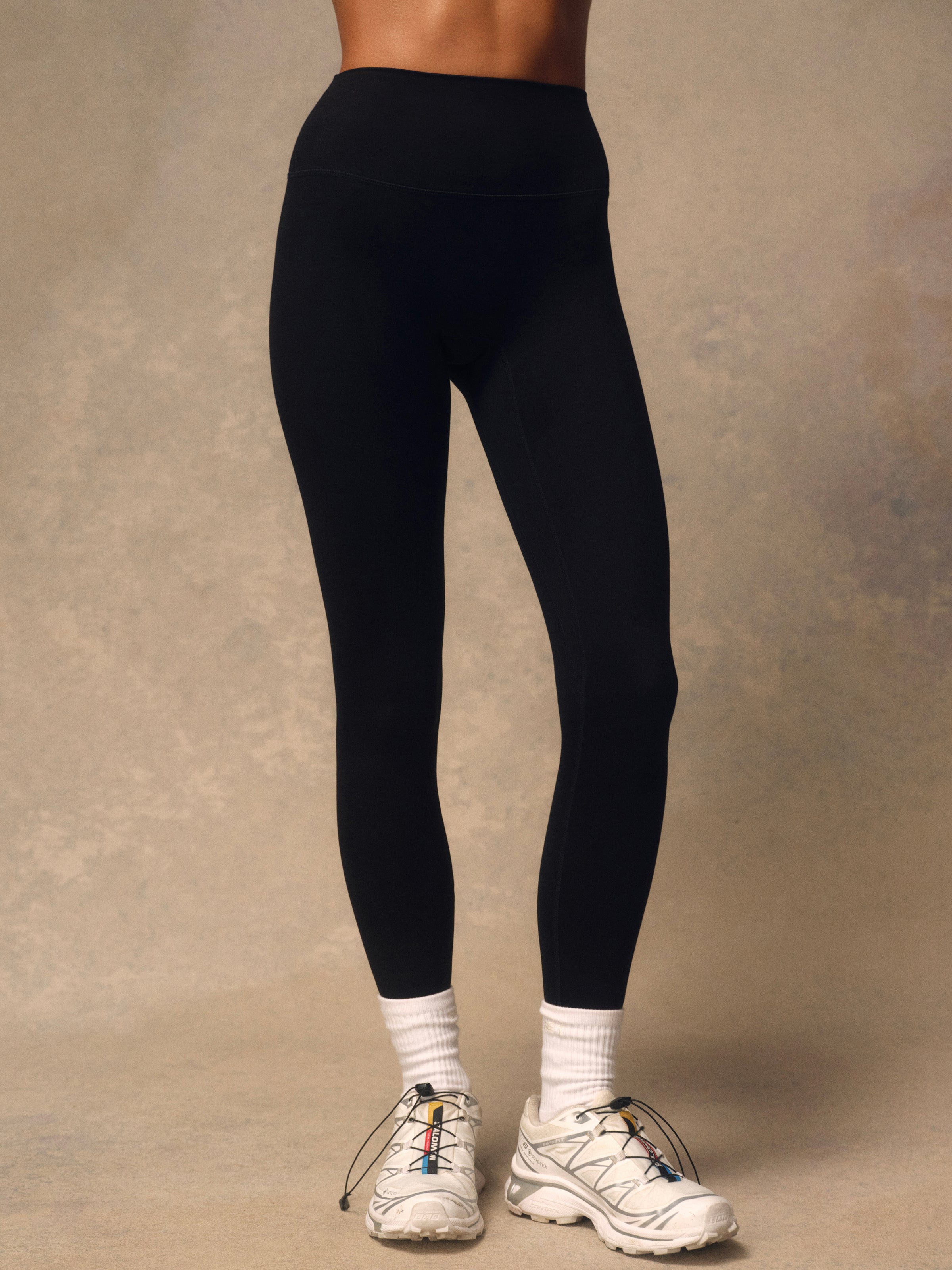Black high-waisted leggings worn by a model, paired with white crew socks and white trail running shoes, shown from the waist down against a textured beige studio backdrop.