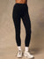 Black high-waisted leggings worn by a model, paired with white crew socks and white trail running shoes, shown from the waist down against a textured beige studio backdrop.
