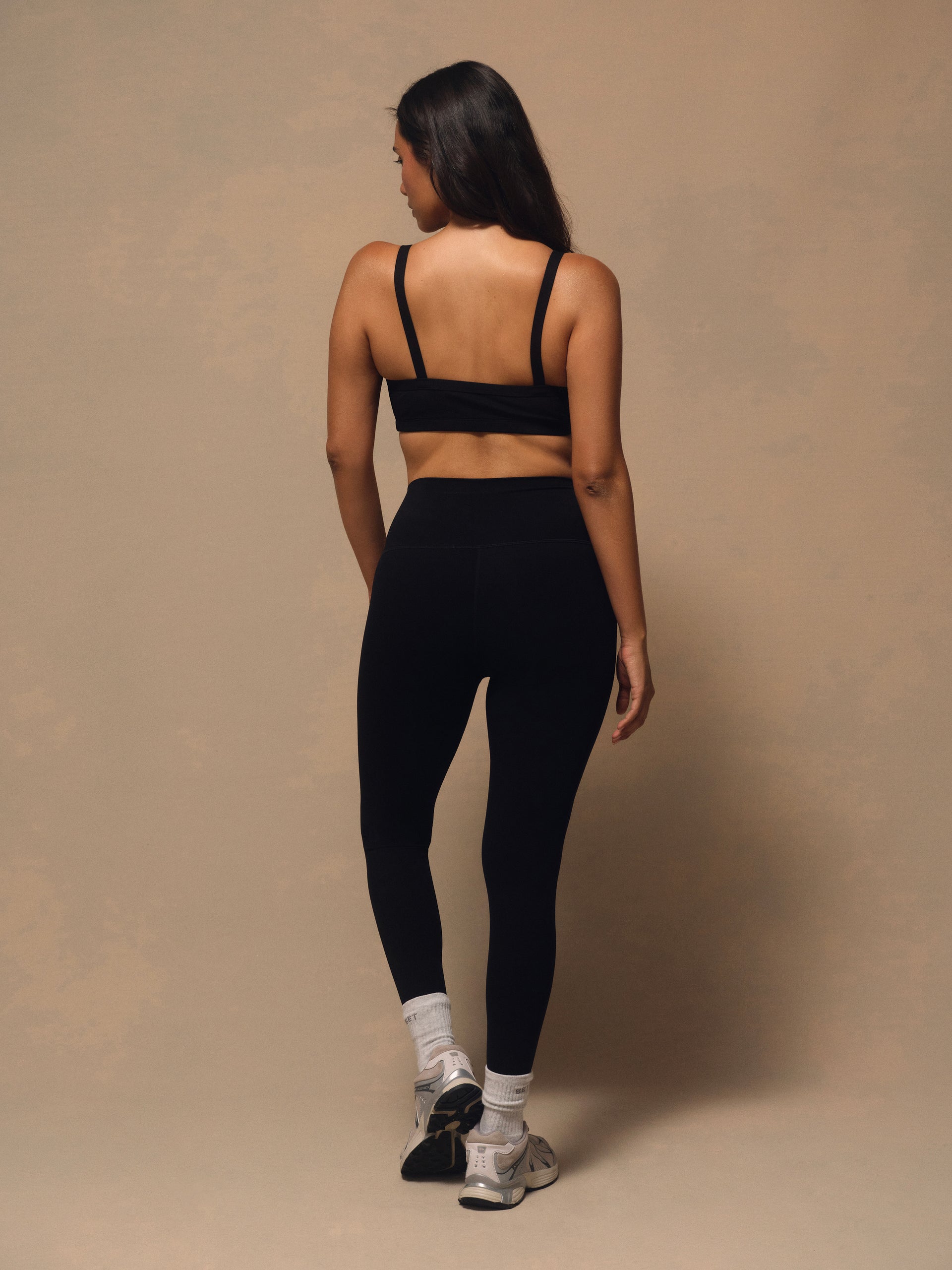 A woman is shown from behind wearing a black sports bra and black high-waisted leggings, standing against a neutral beige studio backdrop.
