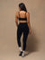 A woman is shown from behind wearing a black sports bra and black high-waisted leggings, standing against a neutral beige studio backdrop.