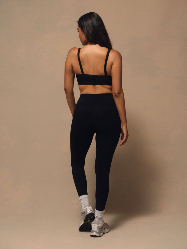 A woman is shown from behind wearing a black sports bra and black high-waisted leggings, standing against a neutral beige studio backdrop.