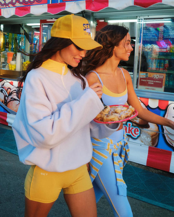 Two women at a carnival, one holding food, with a colorful booth in the background.
