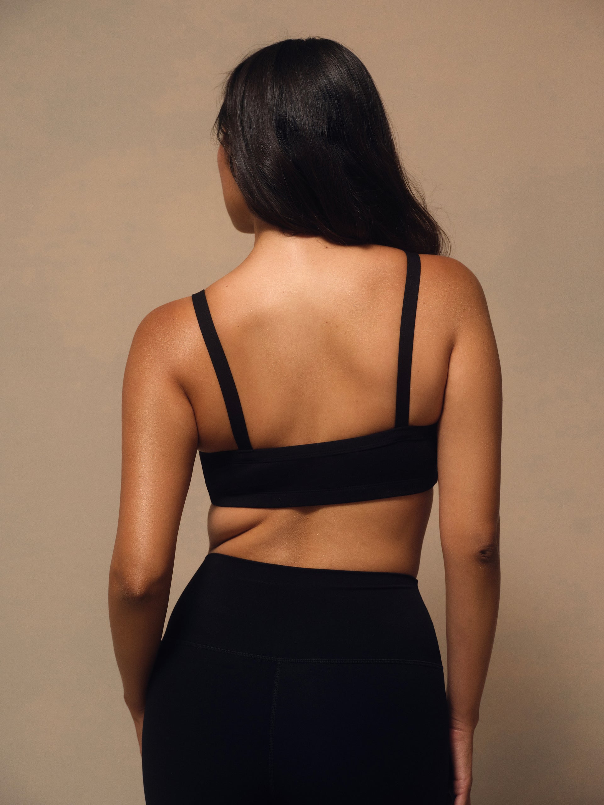 A woman shown from behind wearing a black sports bra with wide shoulder straps and a wide underband, paired with black leggings, against a plain beige studio background.