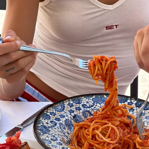 Person eating spaghetti with a fork, wearing a white tank top with 'SET' logo, on a table with a floral plate.