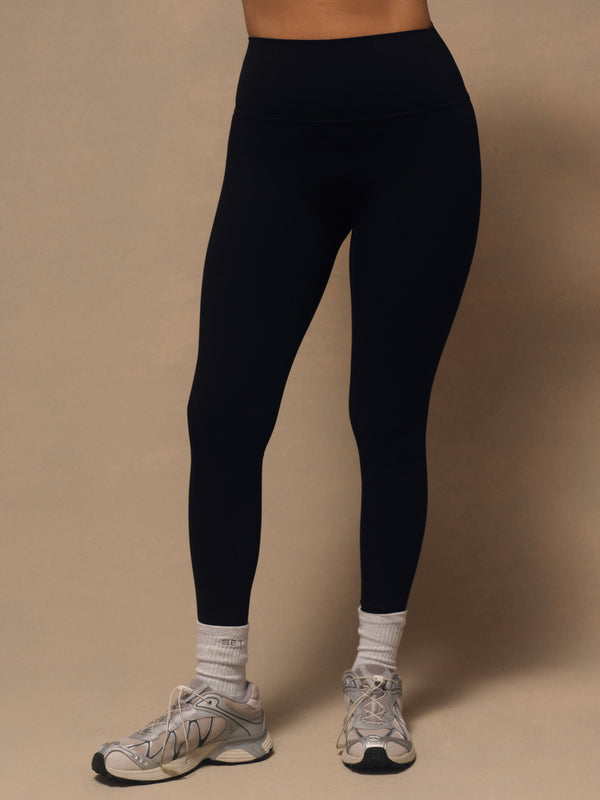 A person wearing black high-waisted leggings, white socks, and gray running shoes, standing against a plain beige studio background.