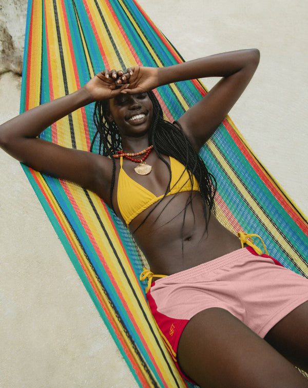A woman reclining in a colorful striped hammock wearing a yellow triangle bikini top, pink bikini bottoms, and red side-tie bottoms, with a neutral outdoor background.