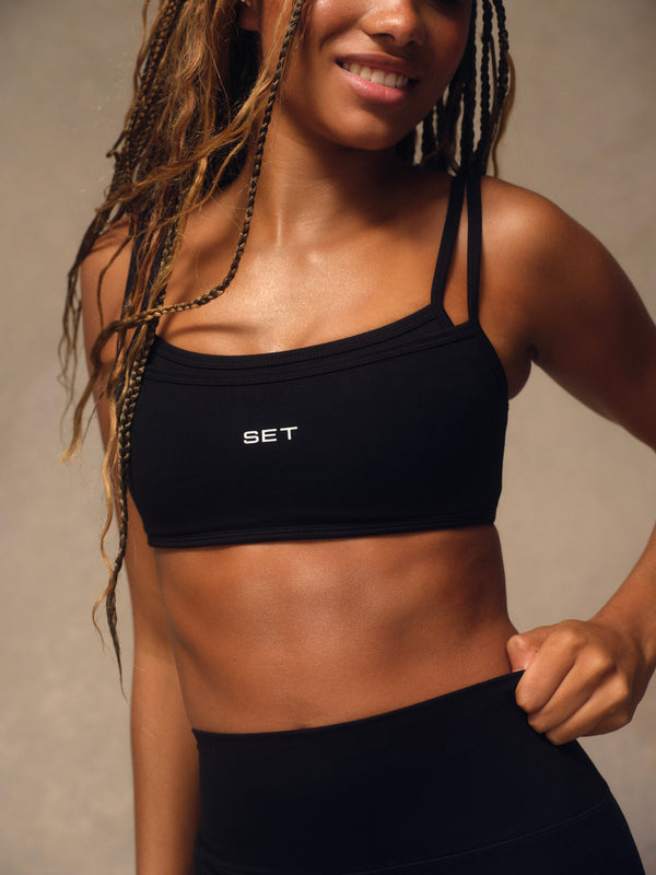A woman wearing a black athletic sports bra with thin straps and a small white 'SET' logo on the front, paired with black athletic bottoms, against a neutral beige background.
