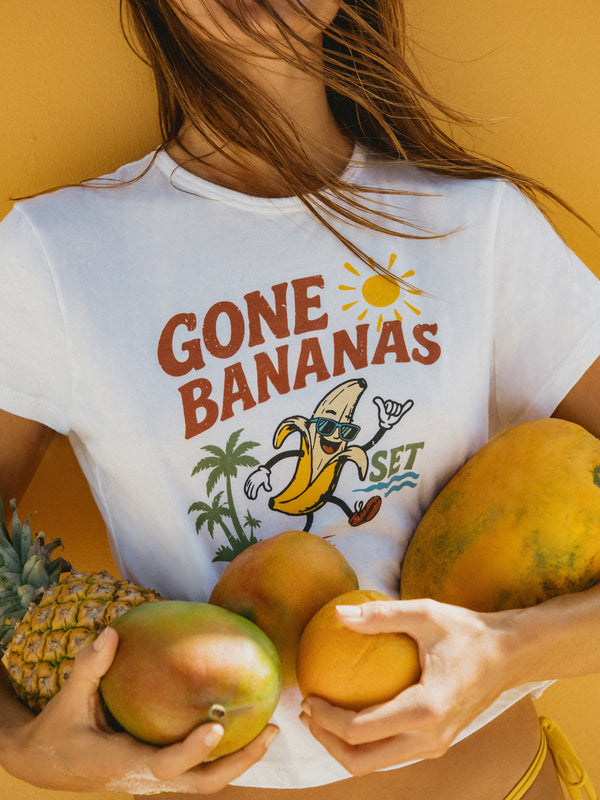 A woman wearing a white graphic t-shirt with the text 'GONE BANANAS SET' and a banana illustration, holding fresh fruits like mangoes and a pineapple against a solid yellow background.