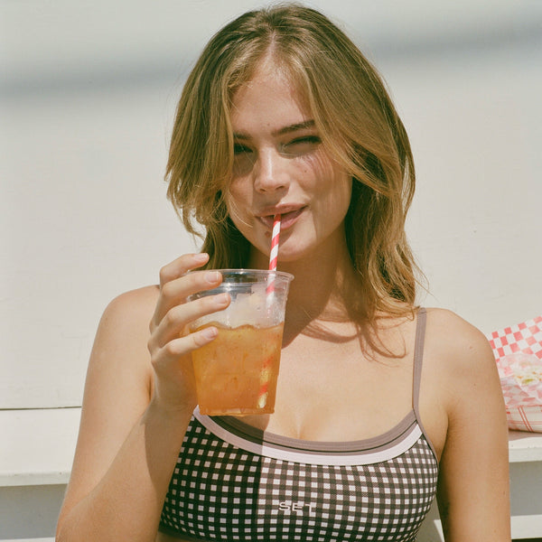 A woman wearing a black and white gingham bikini top and matching bottoms, holding a clear plastic cup with an orange drink and a red-and-white striped straw against a bright white background.