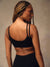 A woman with braided hair is shown from the back wearing a black sports bra with double shoulder straps and black leggings, set against a neutral textured studio background.