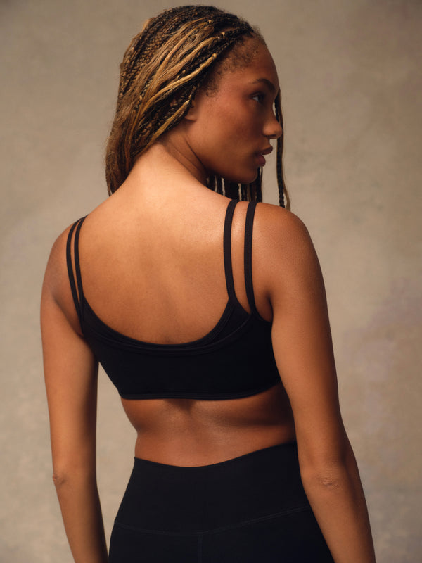 A woman with braided hair is shown from the back wearing a black sports bra with double shoulder straps and black leggings, set against a neutral textured studio background.