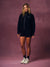 A woman stands against a textured mauve studio backdrop wearing a black zip-up hoodie with a white 'SET' logo on the chest, matching black athletic shorts, black socks, and off-white sneakers.
