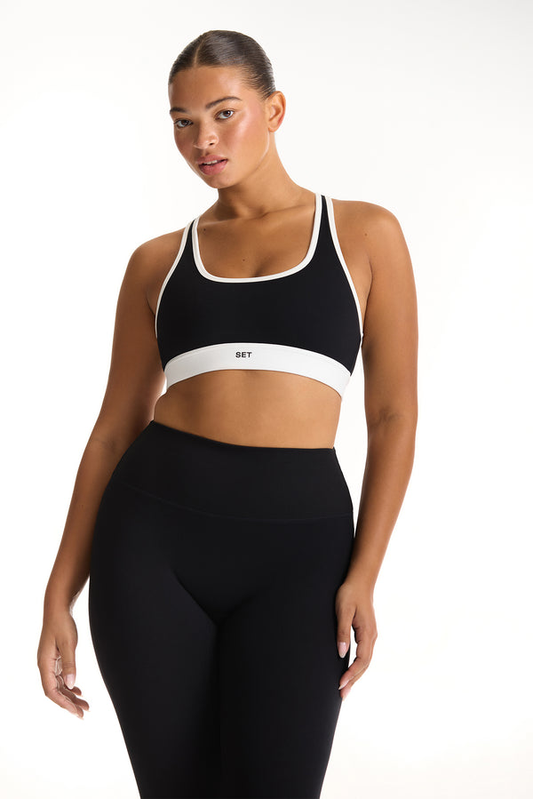 Woman wearing a black sports bra and leggings with a white brand logo on a white background 