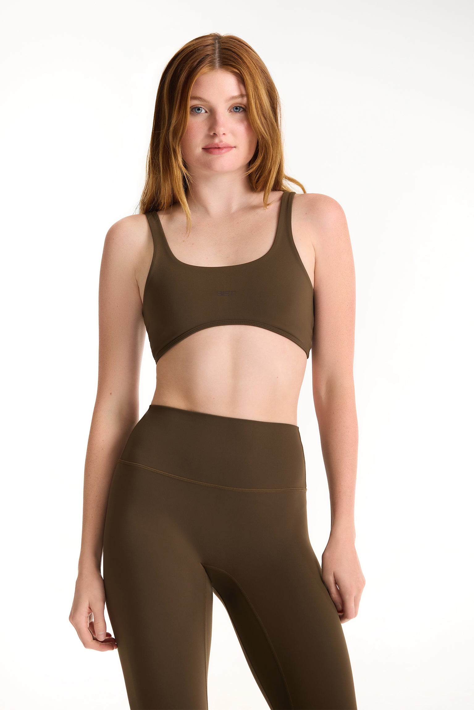 Woman wearing a brown sports bra and leggings on a white background <model-info>Dickey wears a size S and is 5’9”, 24” waist, 32” bust.</model-info>