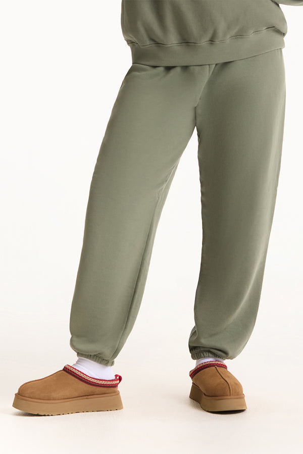 Person wearing olive green sweatpants and brown slippers on a white background 