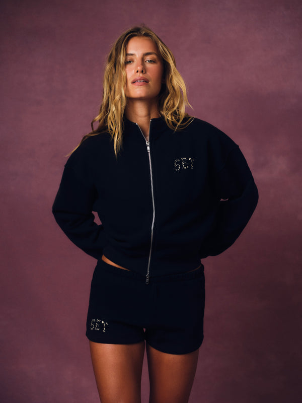 A woman modeling a black zip-up sweatshirt and matching black shorts with silver 'SET' lettering on a textured mauve background. 