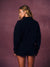 A woman with blonde hair is shown from behind wearing a black crewneck sweatshirt and matching black shorts, standing against a textured mauve-pink studio backdrop.
