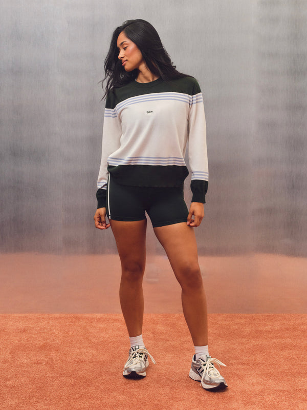 A woman modeling a white, black, and navy striped long-sleeve crewneck sweatshirt with matching black athletic shorts, white socks, and white sneakers, standing on a terracotta-colored surface against a textured gray background. 