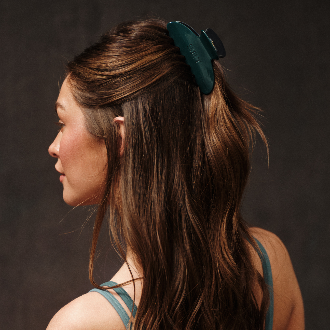 Woman with long brown hair wearing a green hair clip against a dark background