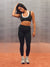 A woman modeling a matching dark green activewear set consisting of a sports bra with a white underband and high-waisted leggings with white side stripes, standing on a terracotta-colored surface against a textured gray studio  <model-info>Alana wears a size M and is 5’8”, 28” waist, 35" bust.</model-info>backdrop.