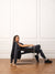 A woman sits on a modern metal-framed chair in a minimalist room with white paneled walls and a wood parquet floor. She is wearing a black sleeveless top, black leggings, and black sneakers, with a black jacket draped over the back of the chair. <model-info>Leah wears a size M and is 5’7”, 25” waist, 32D bust.</model-info>