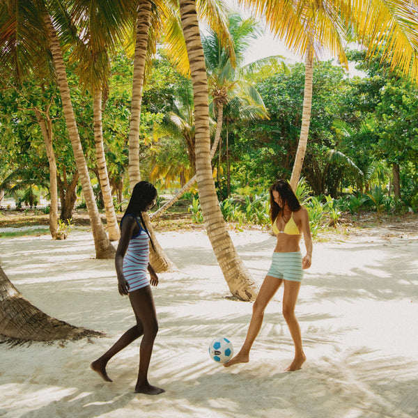 Two women in swimwear play with a soccer ball on a sandy beach under tall palm trees.