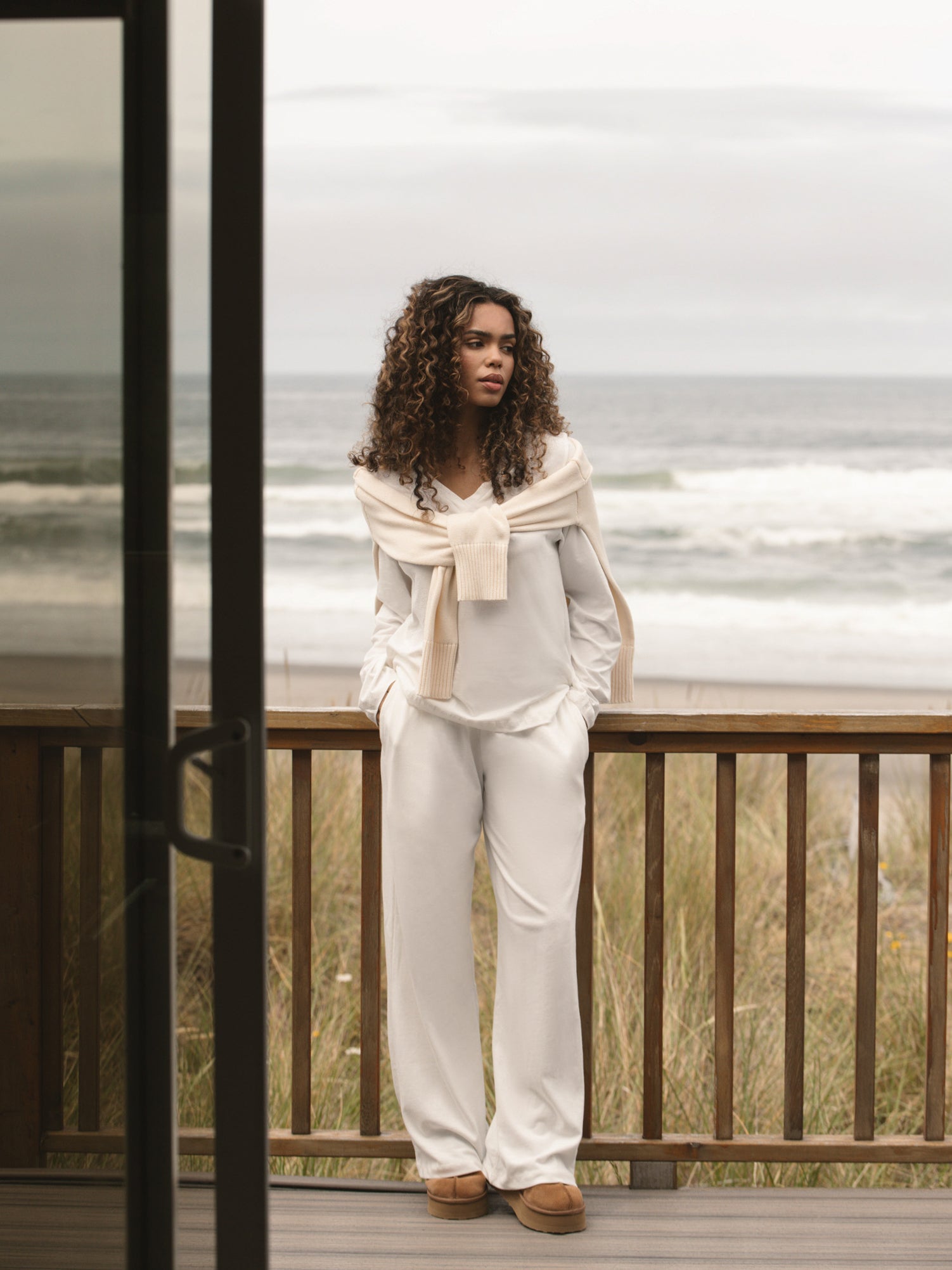Woman standing on a wooden deck by the ocean, wearing a light-colored outfit.