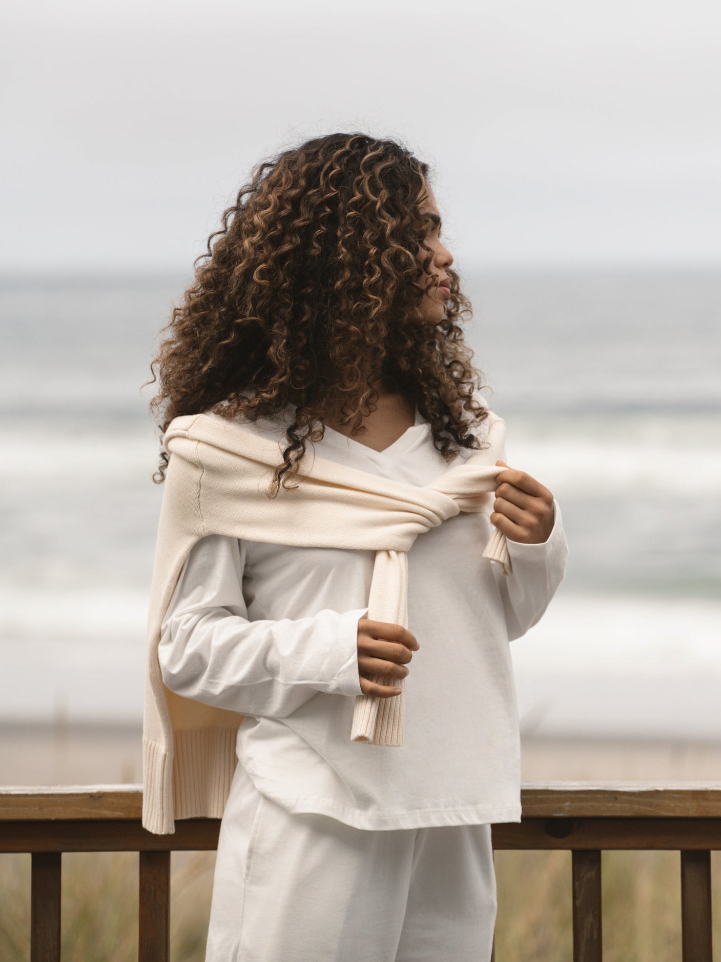 Woman with curly hair wearing a sweater over white outfit against a cloudy sky.