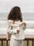 Woman with curly hair wearing a sweater over white outfit against a cloudy sky.