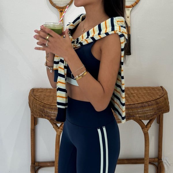 A woman wearing a navy blue activewear set consisting of a sports bra and high-waisted leggings with white side stripes, styled with a striped knit top draped over her shoulders, standing in front of a wicker table.