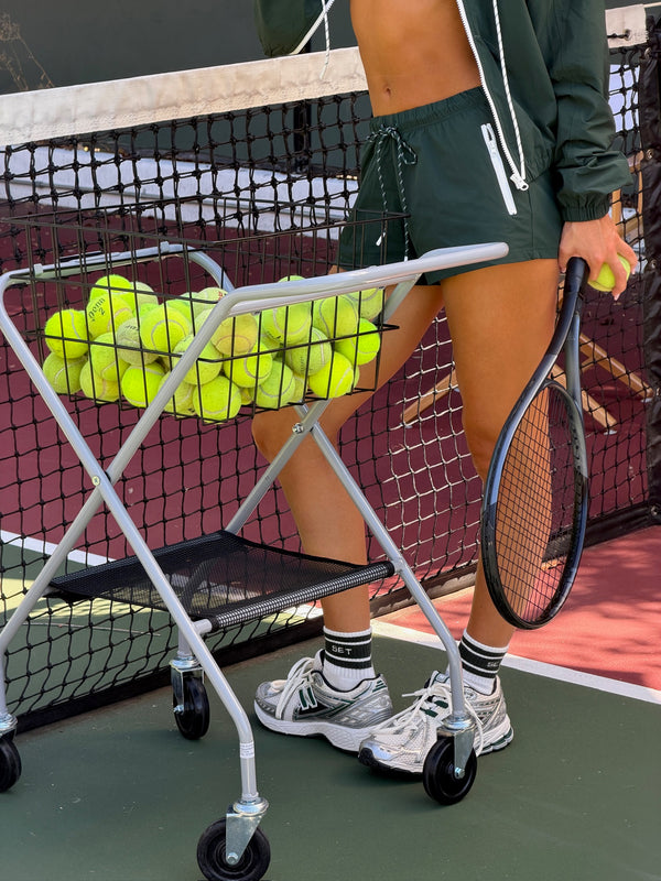 A person on a tennis court wearing a dark green athletic zip-up jacket and matching shorts, standing beside a metal cart filled with yellow tennis balls and holding a tennis racket.