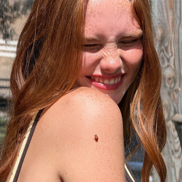 A close-up lifestyle shot of a smiling young woman with freckles and long red hair, looking down at a small ladybug resting on her bare shoulder.