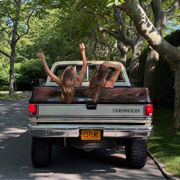 Two women leaning out of the back of a vintage Chevrolet pickup truck parked on a tree-lined road, with their arms raised in the air.