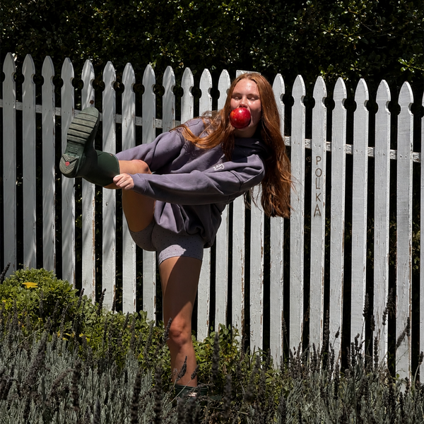 Woman standing in a field putting a pair of boots on while eating an apple.