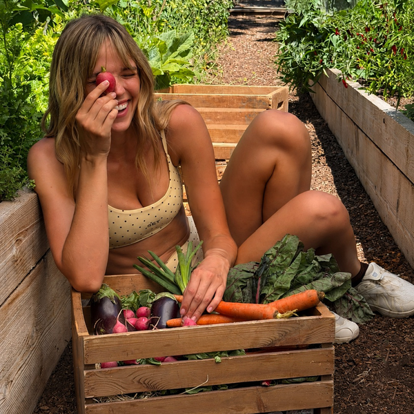 A woman sitting in a garden next to a wooden crate filled with fresh vegetables, including carrots, radishes, and eggplants.