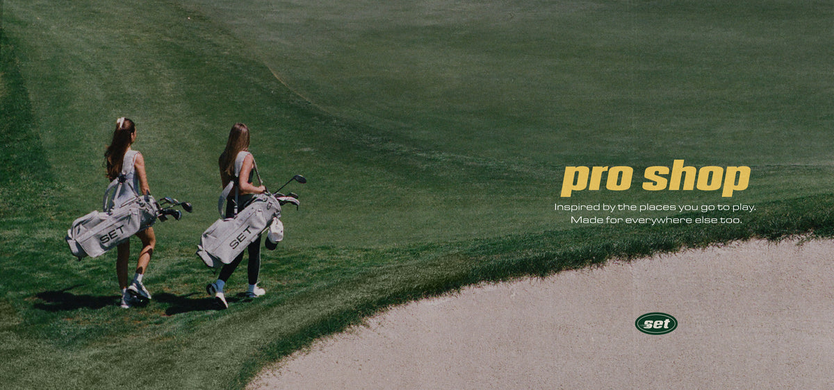 Two women carrying golf bags walk along a grassy golf course beside a sand bunker, with yellow 'pro shop' text and a small SET logo on the right side of the image.