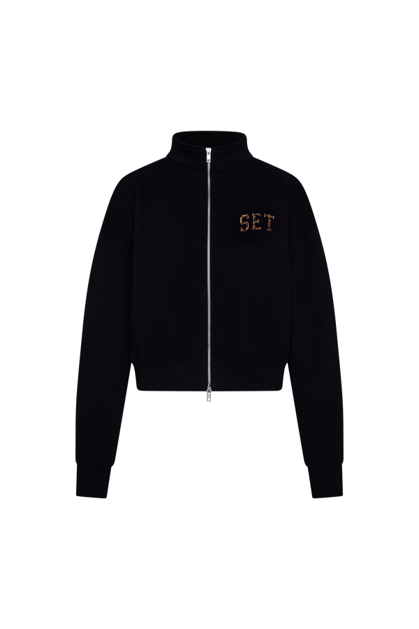 A black cropped zip-up jacket with a stand collar, featuring the letters 'SET' embroidered in gold on the left chest, displayed on a solid white background.