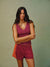 A woman modeling a matching athletic set consisting of a sleeveless V-neck tank top and shorts. The top is a deep berry purple with a small white logo, and the shorts feature orange side panels. <model-info>Jelene wears a size S and is 5’7”, 23” waist, 32AB bust.</model-info>