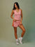 A woman modeling a pink athletic tank top and matching pink shorts with a yellow stripe detail, paired with white socks and sneakers, against a muted sage green studio background.