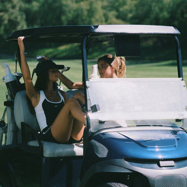 A blue and black golf cart on a golf course path with two people inside, surrounded by green grass and trees under a clear blue sky.